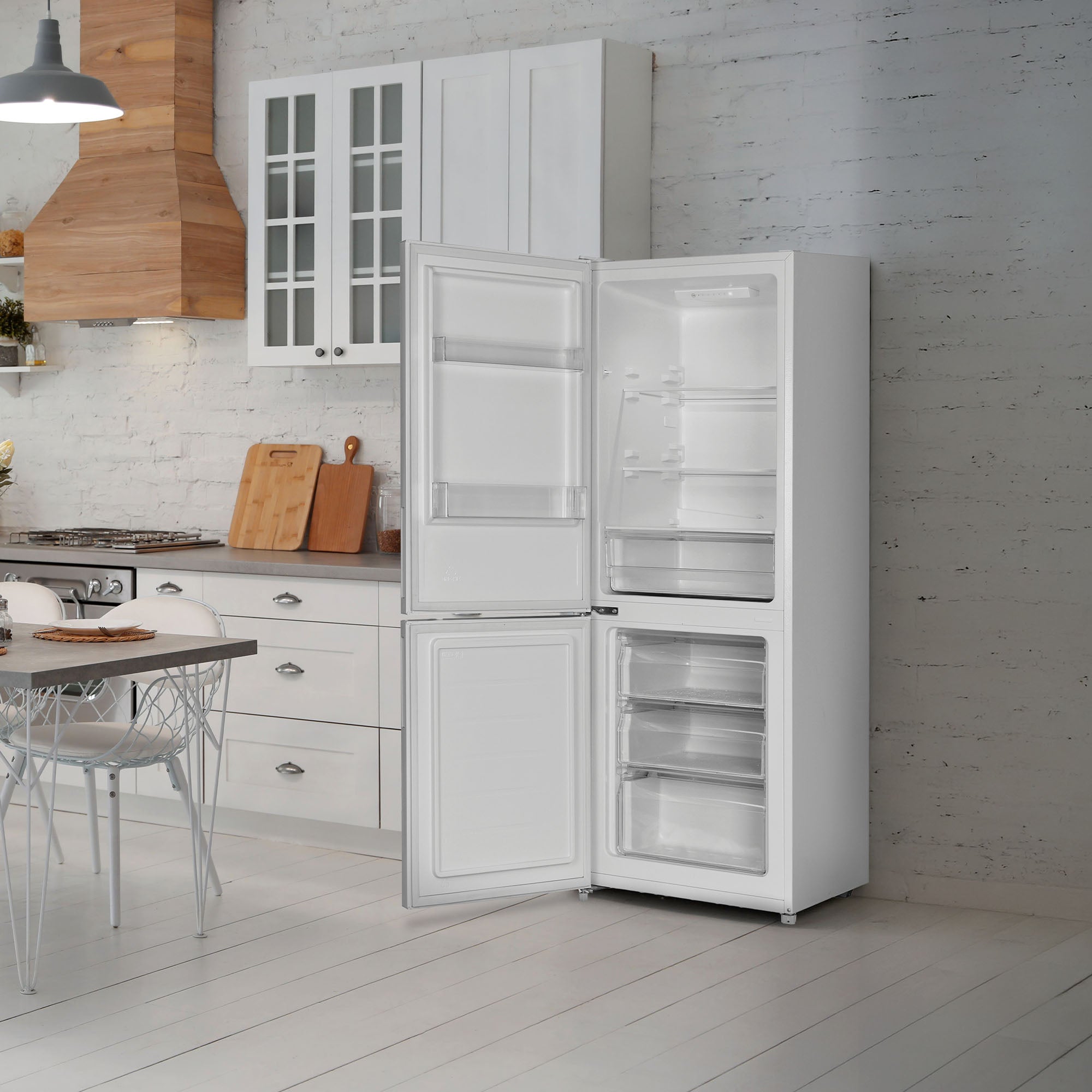 A modern white kitchen features the Statesman v60/40 Low Frost Fridge Freezer from Statesman Appliances, displayed open to reveal its adjustable shelving and spacious drawers. Also visible are sleek white cabinets, a wooden countertop, and a dining table with chairs.