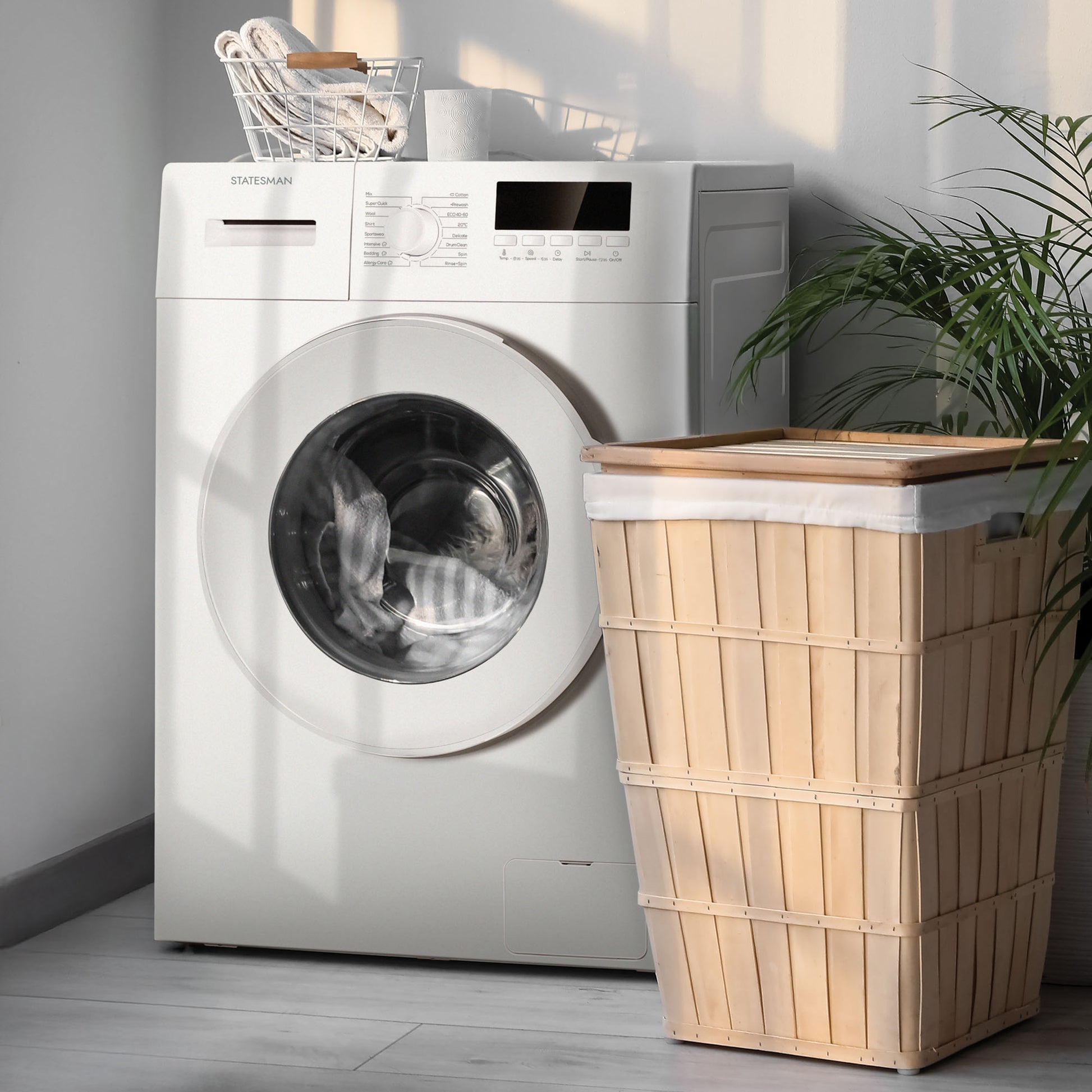 A Statesman Freestanding Washing Machine (FWM0814W, White, 8kg, 1400rpm) stands beside a lidded wooden laundry basket. Nearby, a plant and streaming sunlight create soft shadows on the floor and wall.