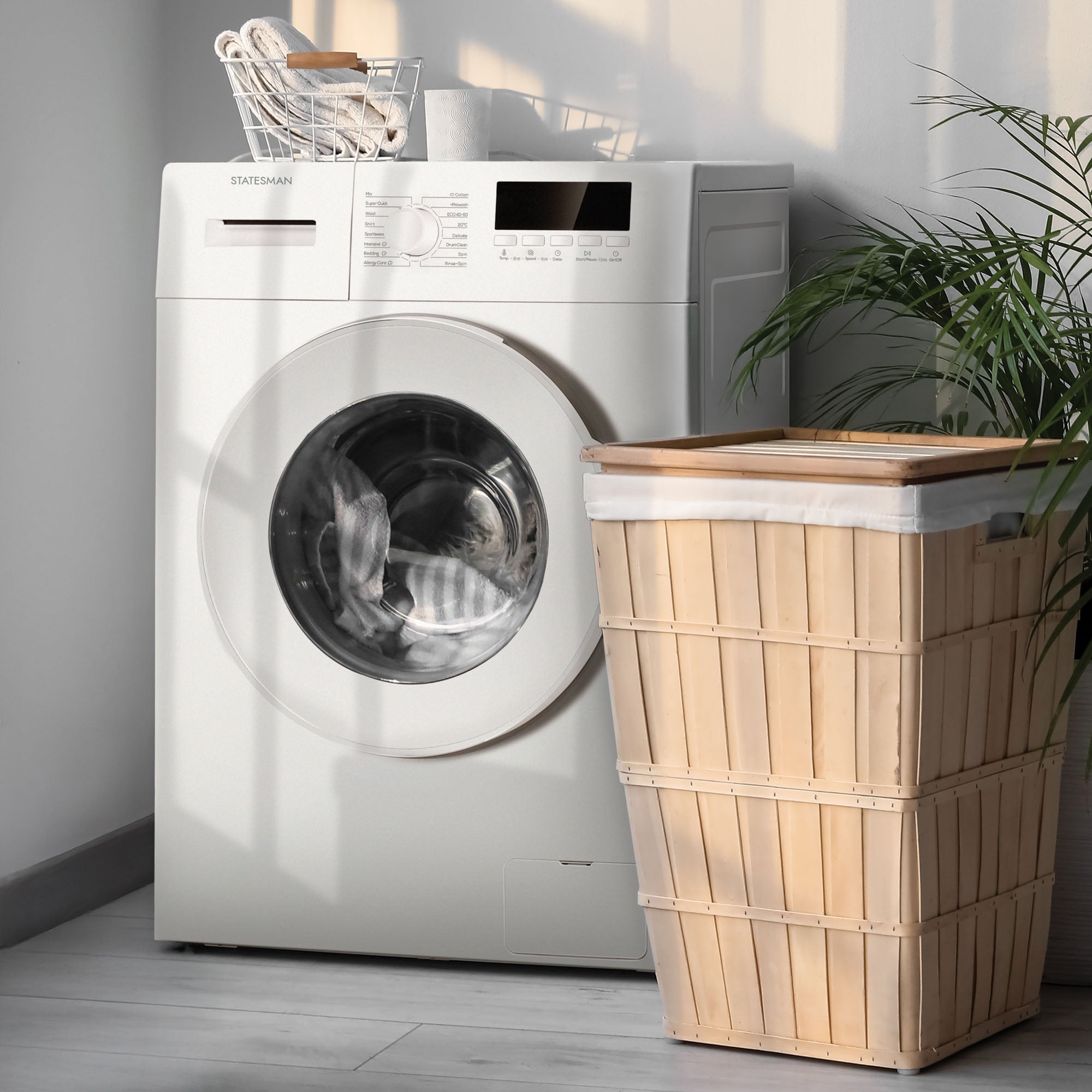A Statesman Freestanding Washing Machine (FWM0814W, White, 8kg, 1400rpm) stands beside a lidded wooden laundry basket. Nearby, a plant and streaming sunlight create soft shadows on the floor and wall.