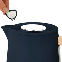 A hand removes a small, semi-circular metal filter from the spout of the Statesman Appliances 1.7L Scandi Kettle, which features a dark finish, wooden handle, metallic lid, and cordless base.