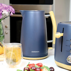 A matte blue Statesman 1.7L Scandi Kettle from Statesman Appliances, featuring a wood-look handle, sits beside a matching toaster on the counter. In front are a glass of tea, grapes, and bread; purple flowers brighten the background.