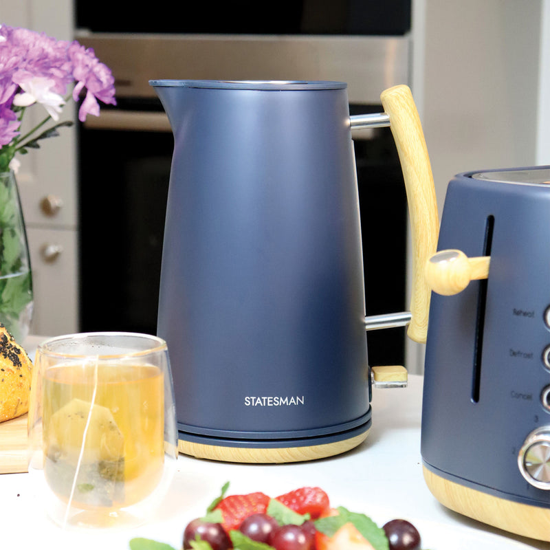 A matte blue Statesman 1.7L Scandi Kettle from Statesman Appliances, featuring a wood-look handle, sits beside a matching toaster on the counter. In front are a glass of tea, grapes, and bread; purple flowers brighten the background.
