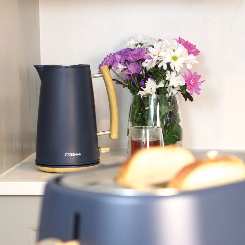 A Statesman Appliances 1.7L Scandi Kettle in navy blue with a wooden handle rests on the kitchen counter beside a vase of colorful flowers, its cordless base keeping it secure while toasted bread is seen in a toaster nearby.