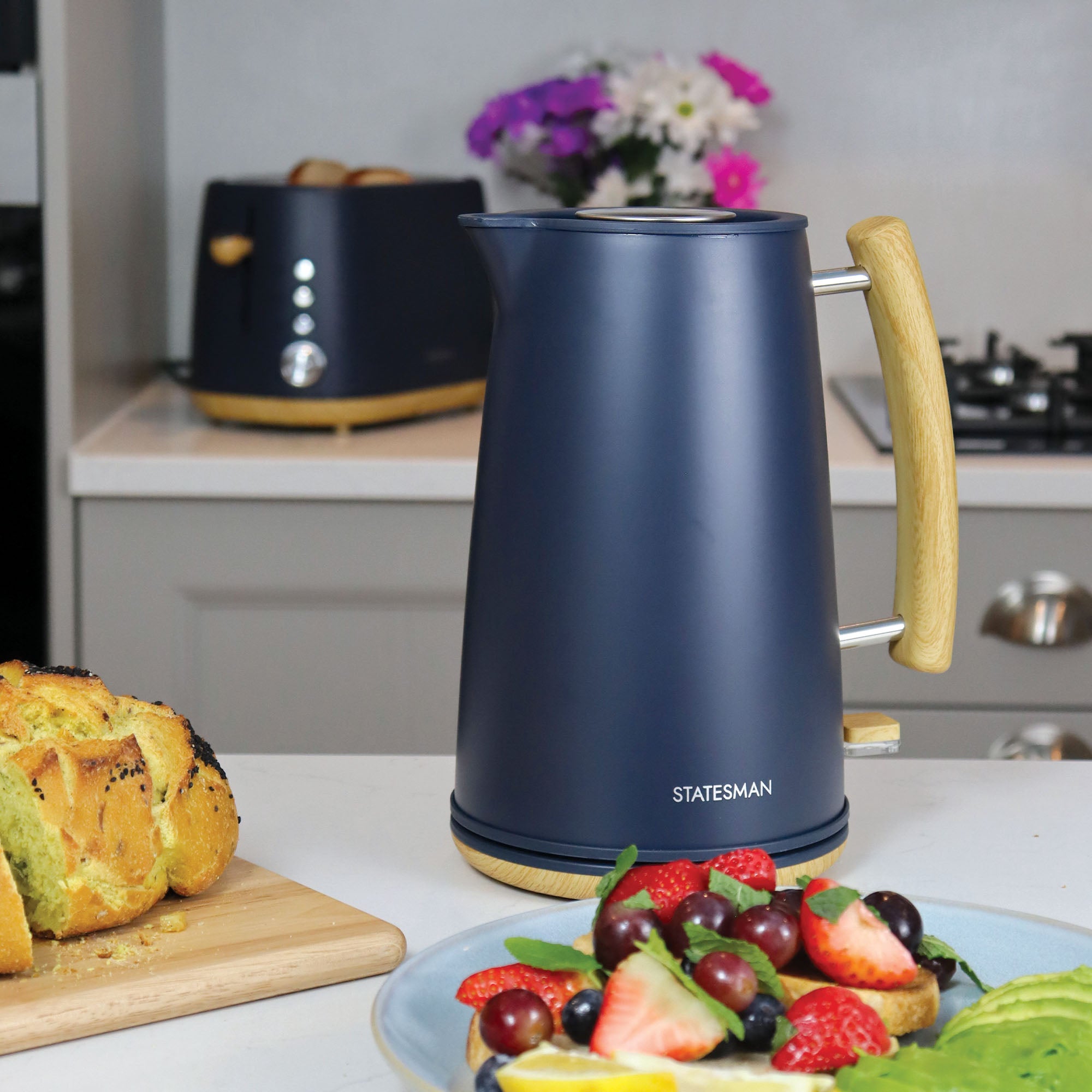 A Statesman Appliances Statesman 1.7L Scandi Kettle in navy blue with wooden accents sits on a kitchen counter near fresh fruit and toast. Its cordless base allows easy movement, with a toaster, sliced bread, and flowers brightening the background.