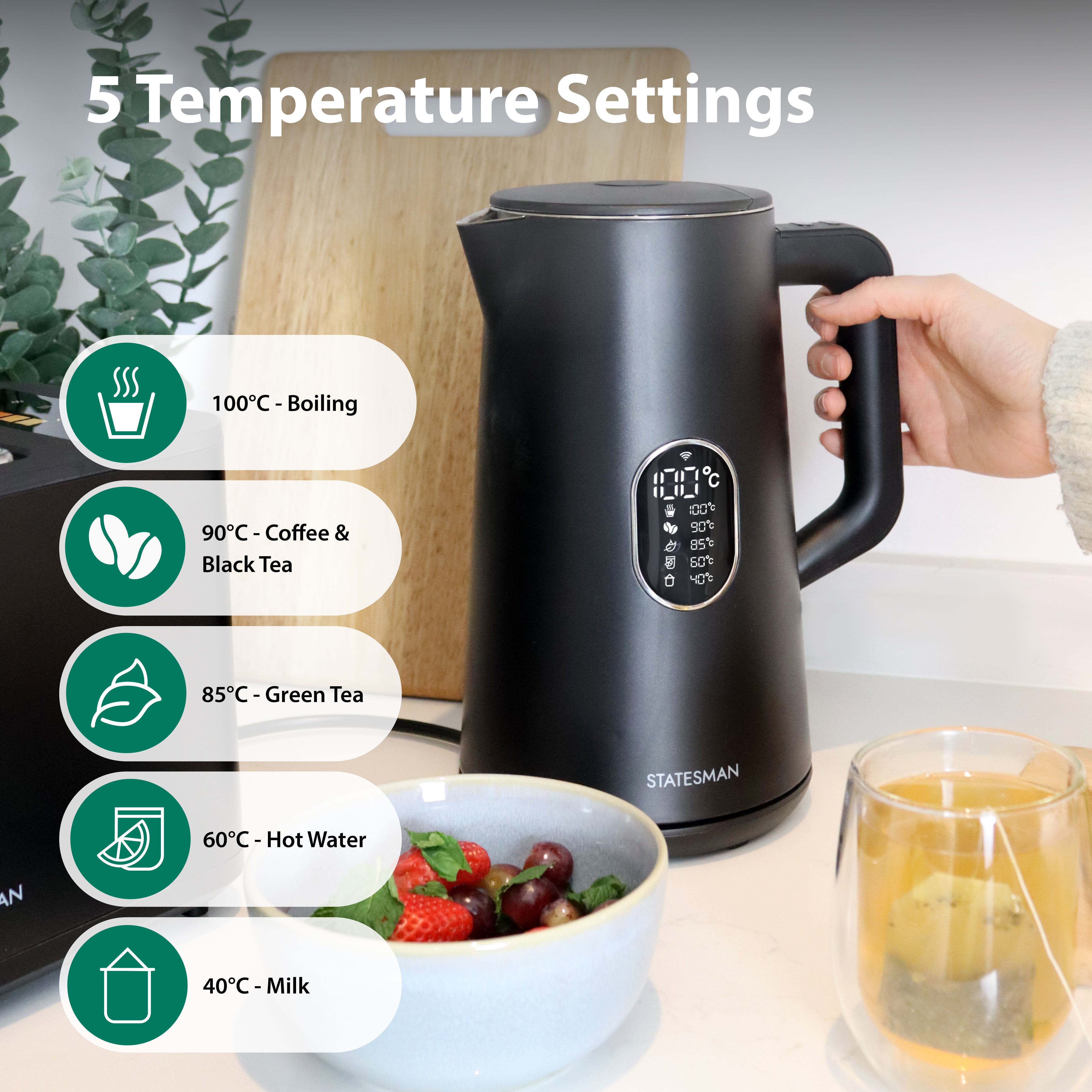 A hand holds the handle of a black Statesman Appliances 1.5L 1800W Smart Jug Kettle with five temperature settings (40°C–100°C) on a kitchen counter, next to strawberries and a glass mug with a hot drink.