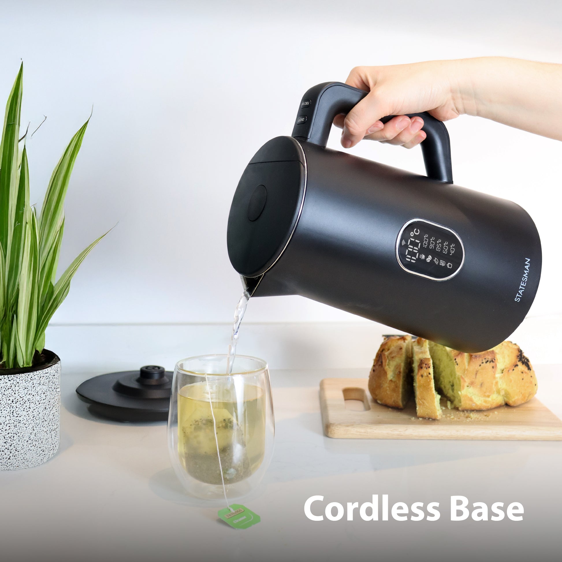 A person pours hot water from a Statesman 1.5L 1800W Smart Jug Kettle, Black by Statesman Appliances into a tea glass. The cordless base is shown near a plant and sliced bread on a wooden cutting board, with the text "Cordless Base" in the corner.