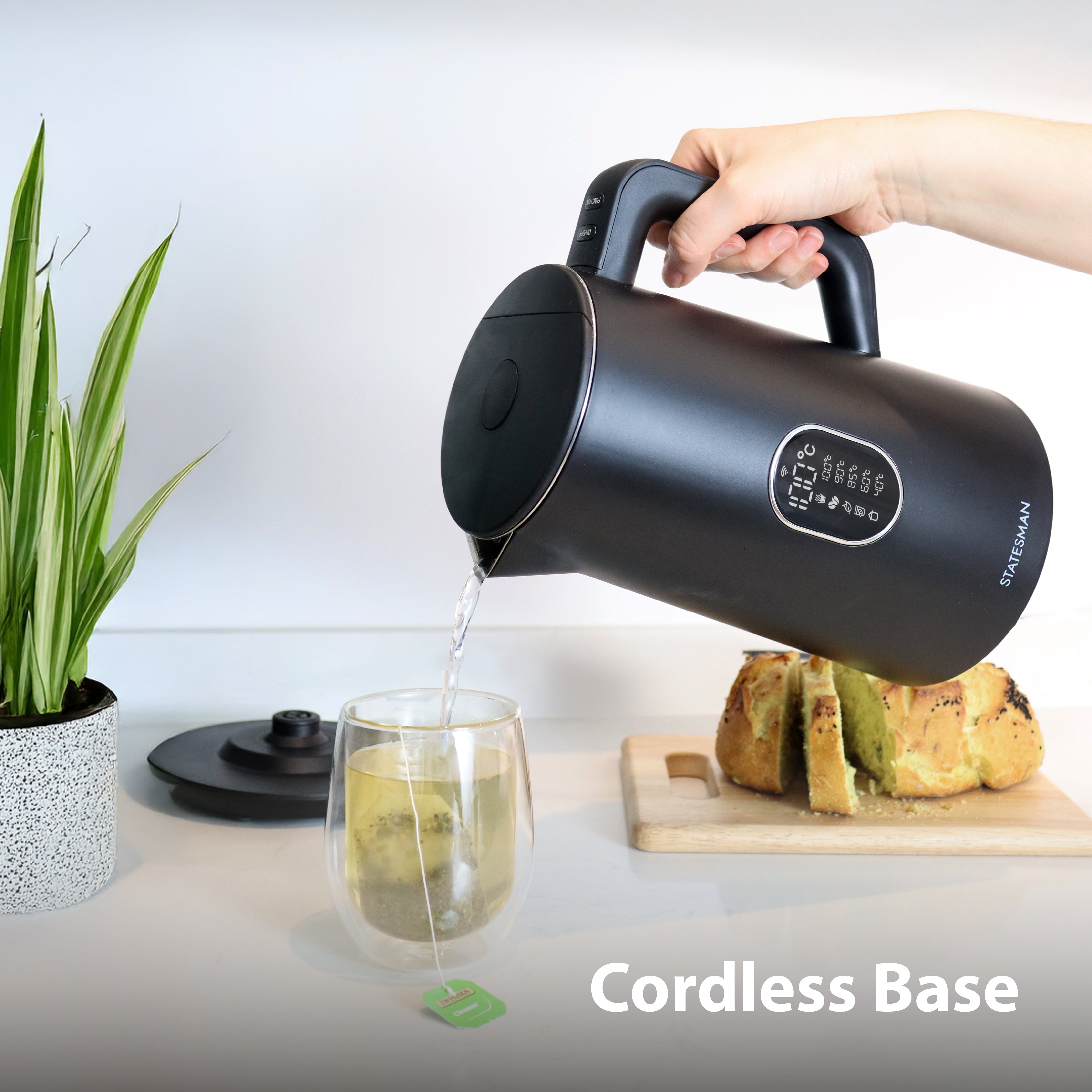 A person pours hot water from a Statesman 1.5L 1800W Smart Jug Kettle, Black by Statesman Appliances into a tea glass. The cordless base is shown near a plant and sliced bread on a wooden cutting board, with the text "Cordless Base" in the corner.
