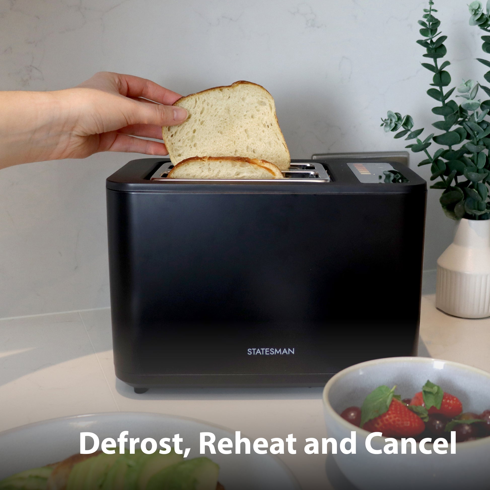 A hand places bread in the Statesman Appliances 900W 2 Slice Digital Toaster, Black, on a kitchen counter. Nearby are strawberries and a plant. On-screen text: Defrost, Reheat and Cancel.