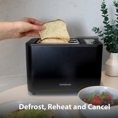 A hand places bread in the Statesman Appliances 900W 2 Slice Digital Toaster, Black, on a kitchen counter. Nearby are strawberries and a plant. On-screen text: Defrost, Reheat and Cancel.