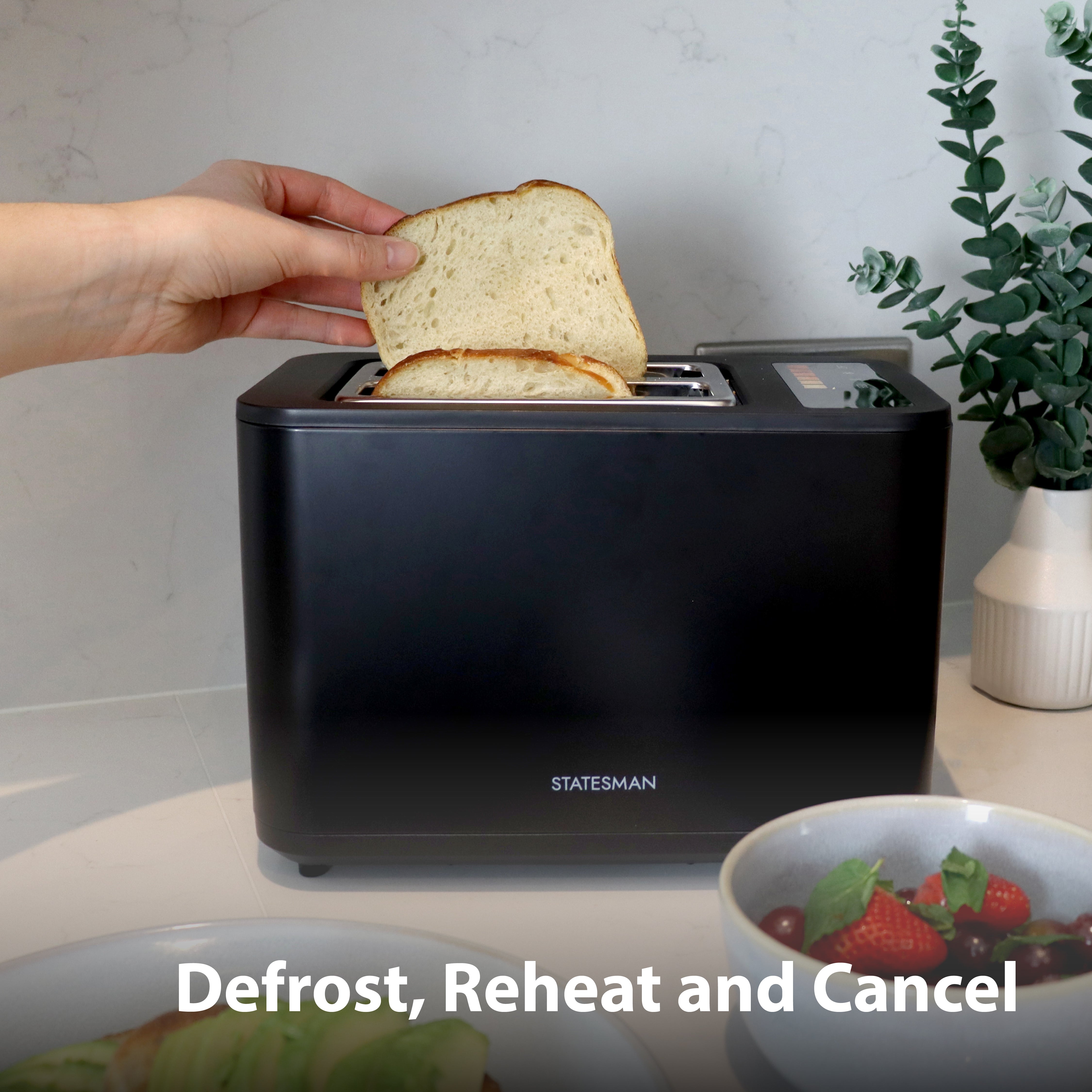 A hand places bread in the Statesman Appliances 900W 2 Slice Digital Toaster, Black, on a kitchen counter. Nearby are strawberries and a plant. On-screen text: Defrost, Reheat and Cancel.