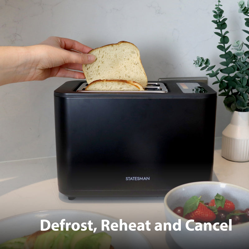 A hand places bread in the Statesman Appliances 900W 2 Slice Digital Toaster, Black, on a kitchen counter. Nearby are strawberries and a plant. On-screen text: Defrost, Reheat and Cancel.