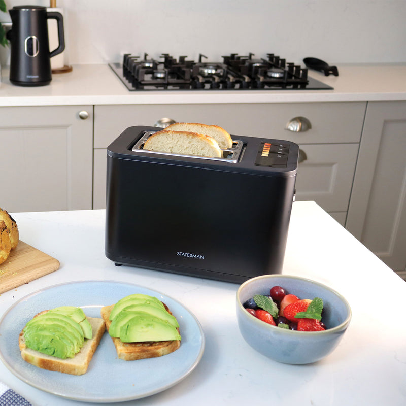 The Statesman 900W 2 Slice Digital Toaster in black from Statesman Appliances sits on a white kitchen counter beside avocado toast, fruit, bread on a cutting board, with a kettle and stove visible in the background.