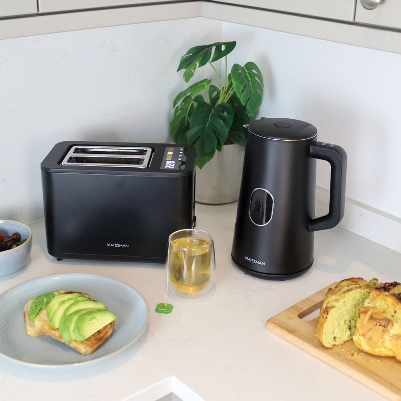 A Statesman Appliances 900W 2 Slice Digital Toaster in black rests on a white counter beside an electric kettle, tea, avocado toast, berries, sliced bread on a wooden board, and a green plant in the background.