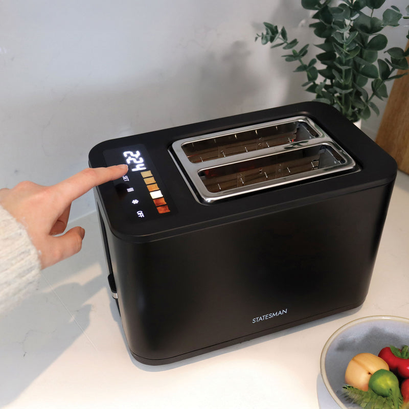 A hand presses a button on the black Statesman Appliances 900W 2 Slice Digital Toaster with wide slots and digital display, placed on a white countertop beside a bowl of vegetables and a green plant.