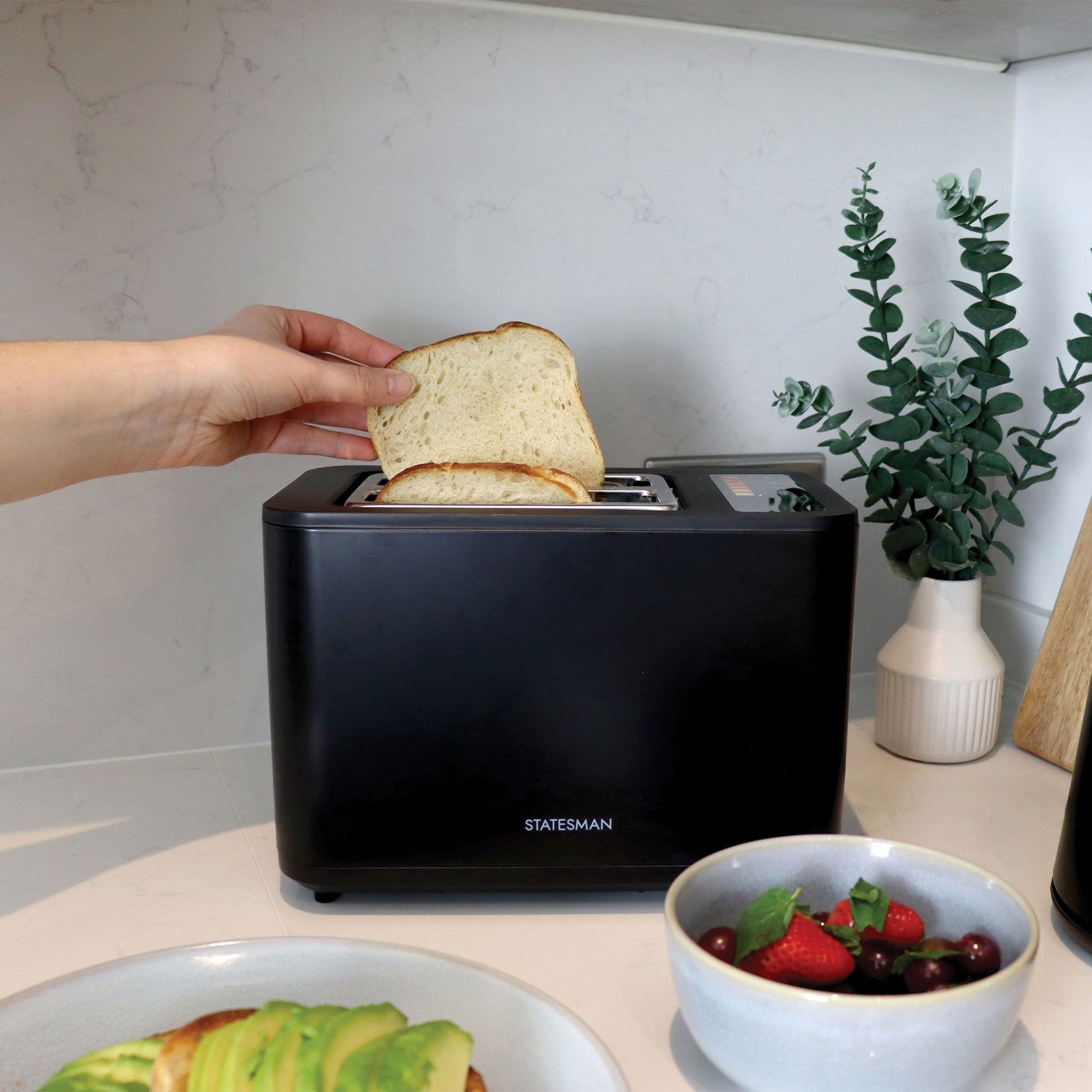 A hand places a slice of bread into the Statesman Appliances 900W 2 Slice Digital Toaster, Black on a kitchen counter, beside avocado toast, fruit, and a vase with green foliage.