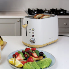 The Statesman 900W 2 Slice Scandi Toaster by Statesman Appliances sits on a kitchen counter, toasting bread in its large slots. In front, a plate displays avocado slices, strawberries, grapes, raspberries, mint leaves, and lemon wedges.