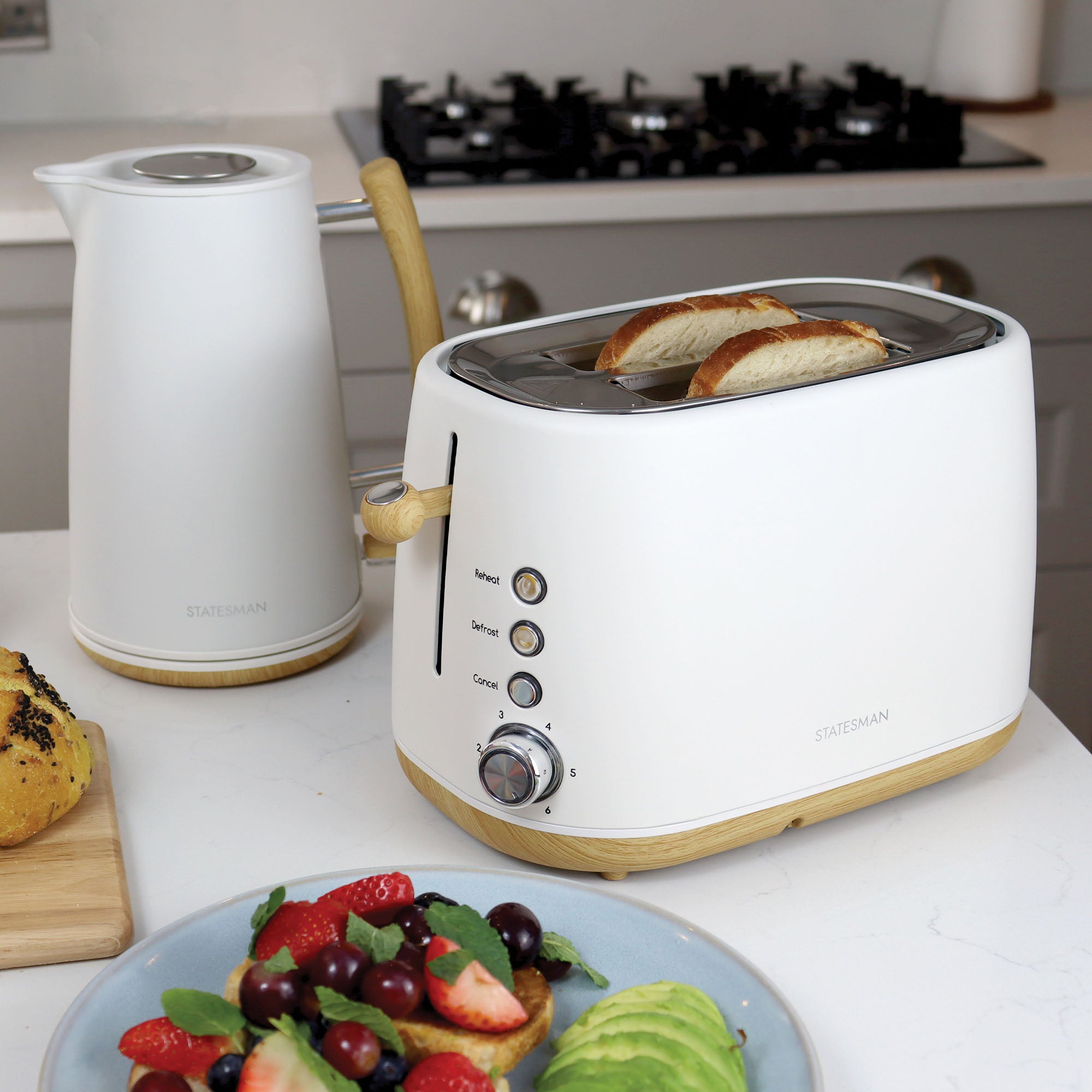 A Statesman Appliances 900W 2 Slice Scandi Toaster sits on a Scandi-style kitchen counter alongside a matching kettle. In the foreground are sliced avocado and fruit on a plate, with a bun on a nearby chopping board.