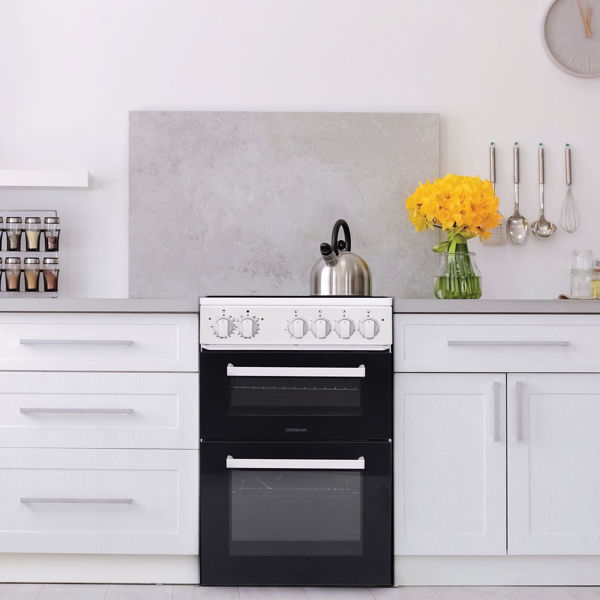 A modern kitchen with white cabinets, a Statesman Twin Cavity Electric Cooker (4 zones, 50cm wide) featuring a separate grill in black and white, a kettle, yellow flowers in a vase, spice jars, utensils hanging on the wall, and a round wall clock.
