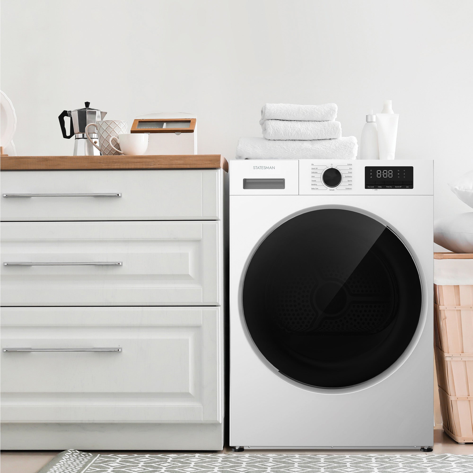 A modern laundry room features a white Statesman Freestanding Heat Pump Tumble Dryer with 8kg load capacity beside a cabinet with three drawers, a wooden countertop holding a coffee maker and towels, and a laundry basket on the right.
